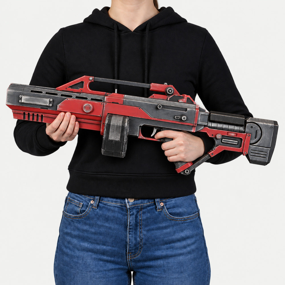 Woman in a black hoodie and blue jeans holding the red, black and gray AR-70 Raptor Nod prop across her upper waist, with a ribbed rectangular twin magazine and skeleton stock, muzzle pointing left.