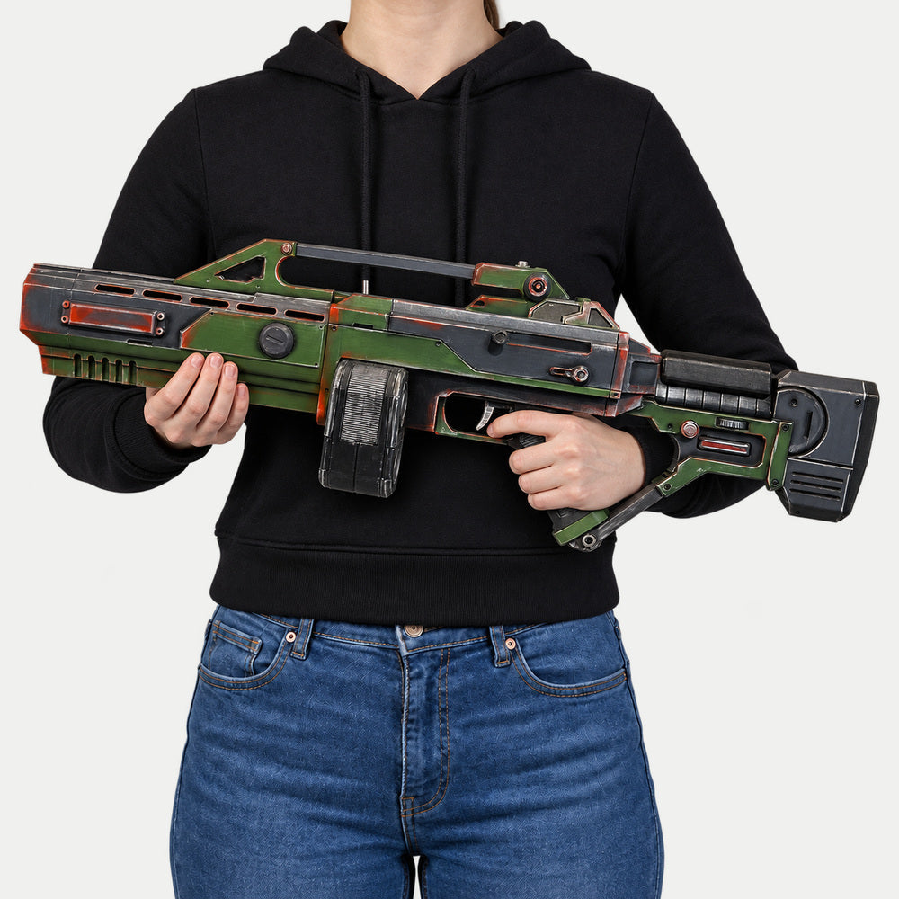 Woman in a black hoodie and jeans holding the olive-green Command & Conquer AR-70 Raptor prop across her body, muzzle facing left.